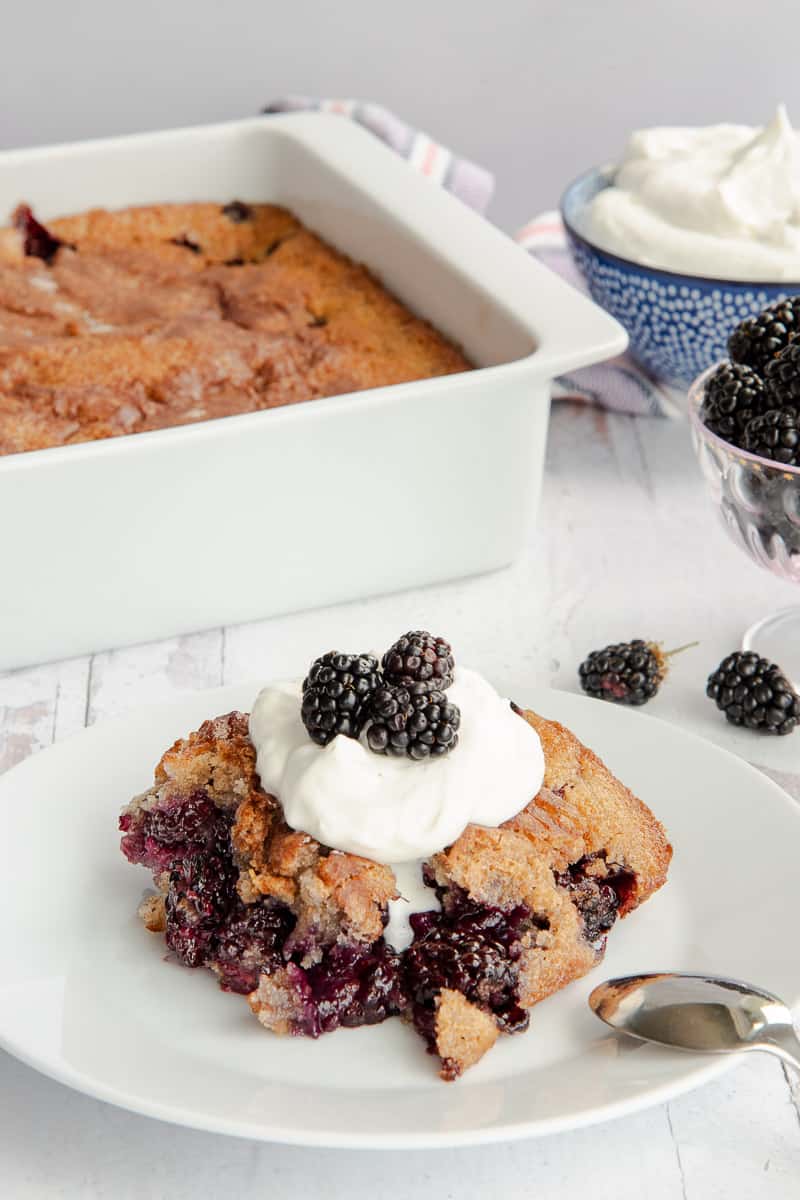 A serving of blackberry cobbler on a white dessert plate garnished with whipped cream and fresh blackberries.