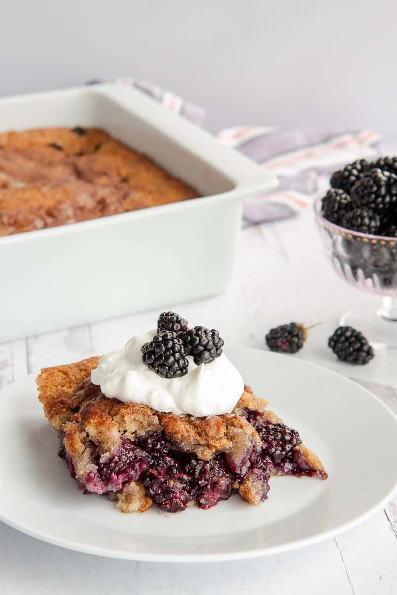 A serving of blackberry cobbler on a white dessert plate in front of a white baking dish with the rest of the cobbler in it.