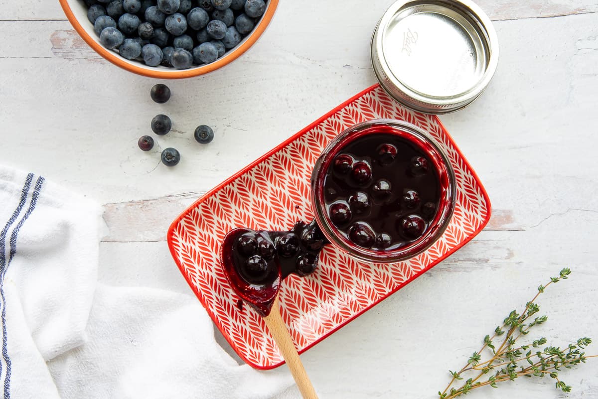 The inside of a glass jar filled with Blueberry Balsamic BBQ Sauce on a orange and white plate next to a bowl of blueberries and a few sprigs of fresh thyme.