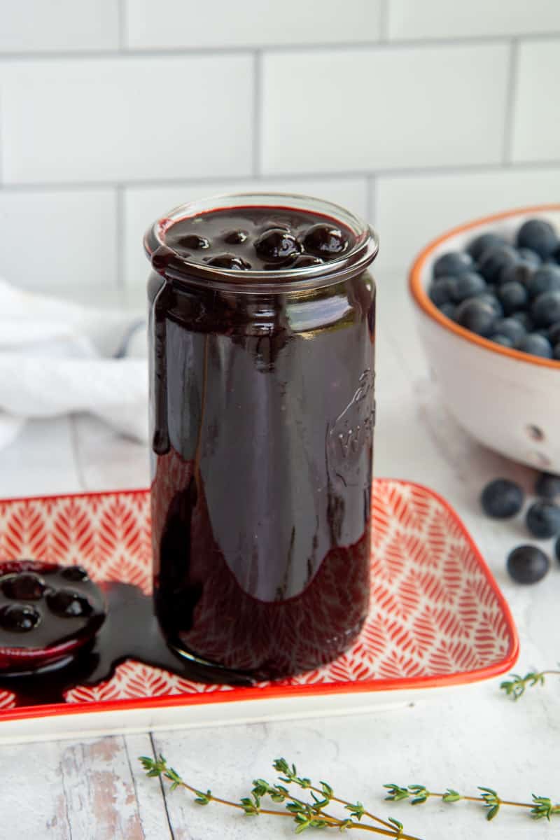 A tall jar of Blueberry Balsamic BBQ Sauce on a orange and white plate next to a white bowl of blueberries.