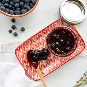 The Blueberry Balsamic BBQ Sauce in a glass jar sits next to a wooden spoonful of sauce on an orange and white plate.