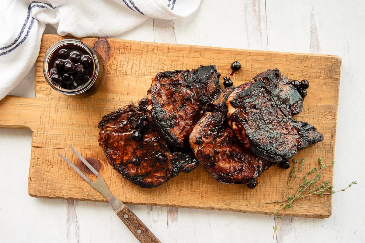 Four Grilled Thick-Cut Pork Chops on a wooden board next to a two-pronged fork and a few sprigs of fresh thyme.