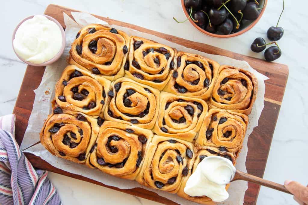 The full batch of Cherry Sweet Rolls on a wooden board as a dollop of almond cream cheese icing is spooned onto one of the rolls.