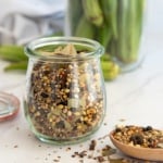 The Flavorful Pickling Spice in a clear glass jar next to a wooden spoonful of the spice blend.