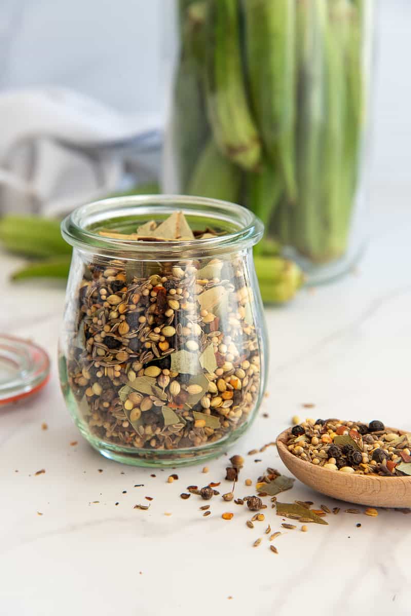 The Flavorful Pickling Spice in a clear glass jar next to a wooden spoonful of the spice blend.