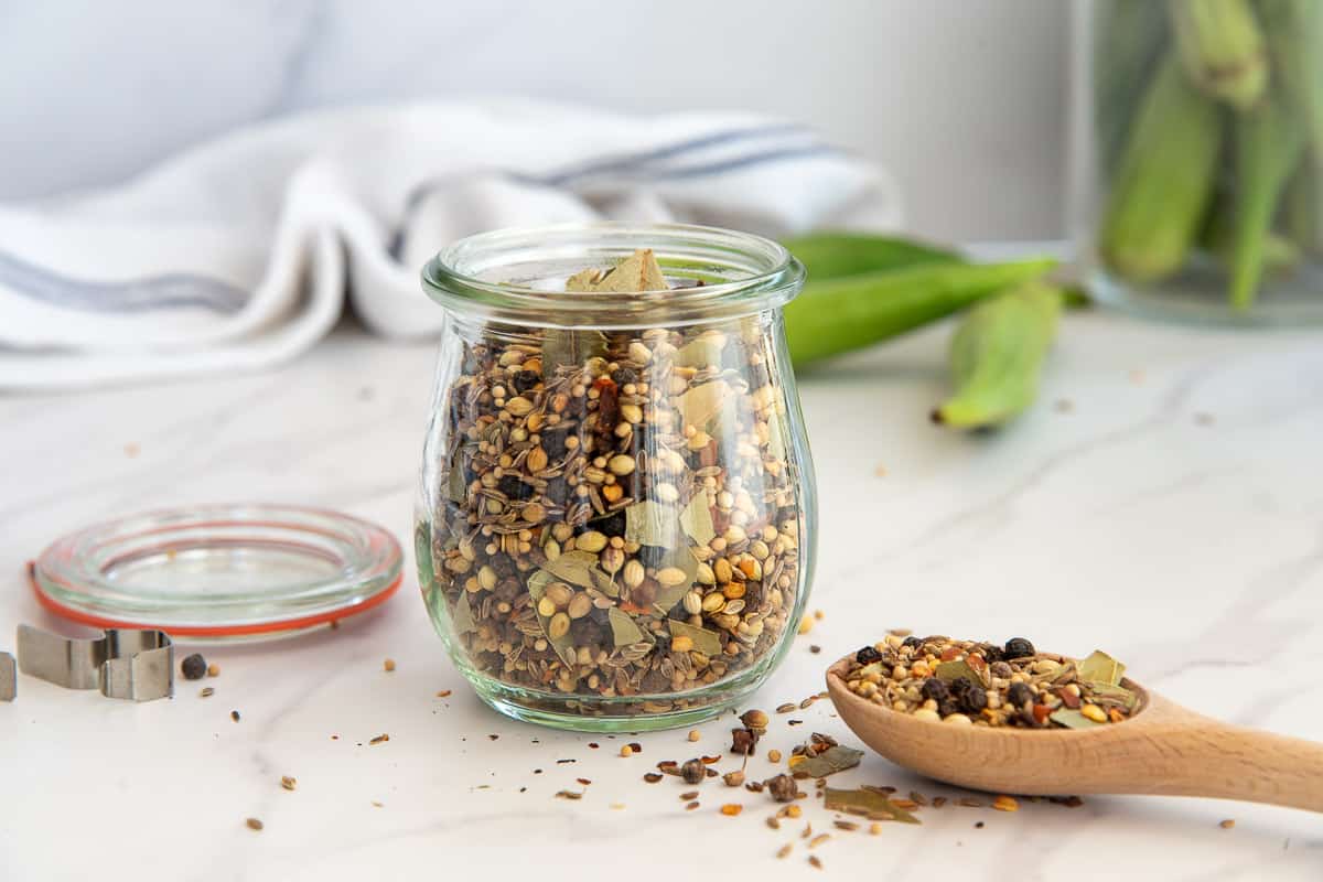 Flavorful Pickling Spice in a clear glass jar next to a wooden spoon with the spice blend on it.
