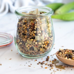 A clear glass jar filled with Flavorful Pickling Spice next to a wooden spoon with the spice blend on it.