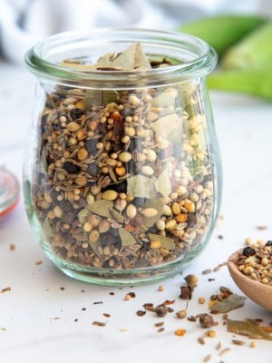 A clear glass jar filled with Flavorful Pickling Spice next to a wooden spoon with the spice blend on it.