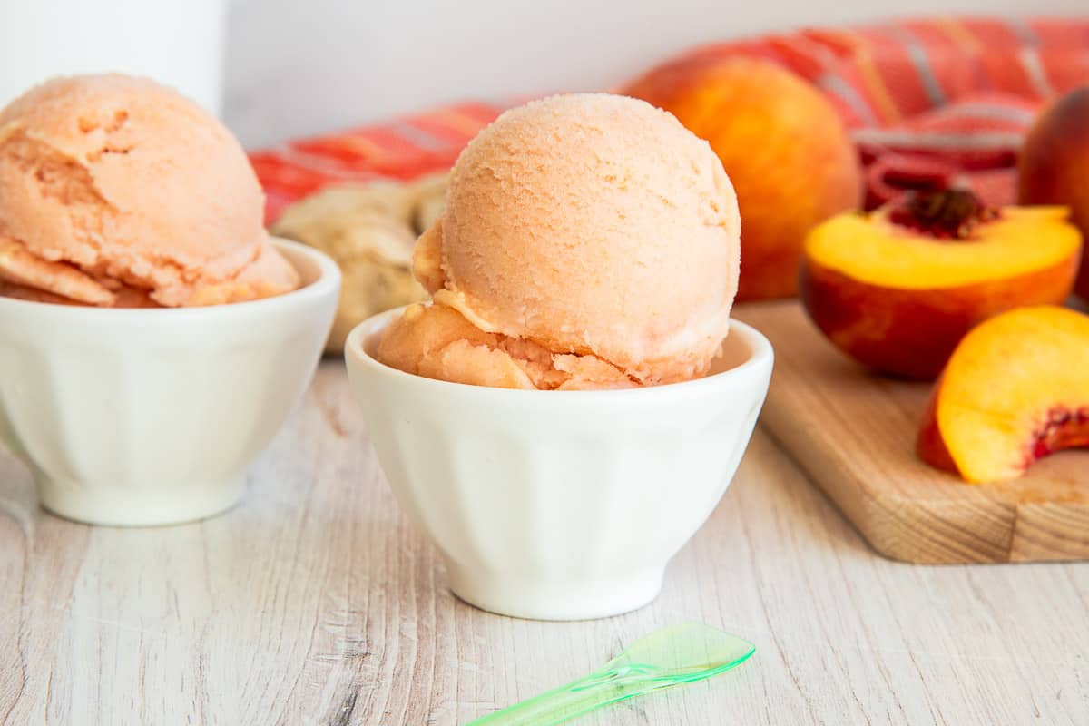 Peach Sorbet in white dessert bowls next to a wooden board with cut peaches on it.