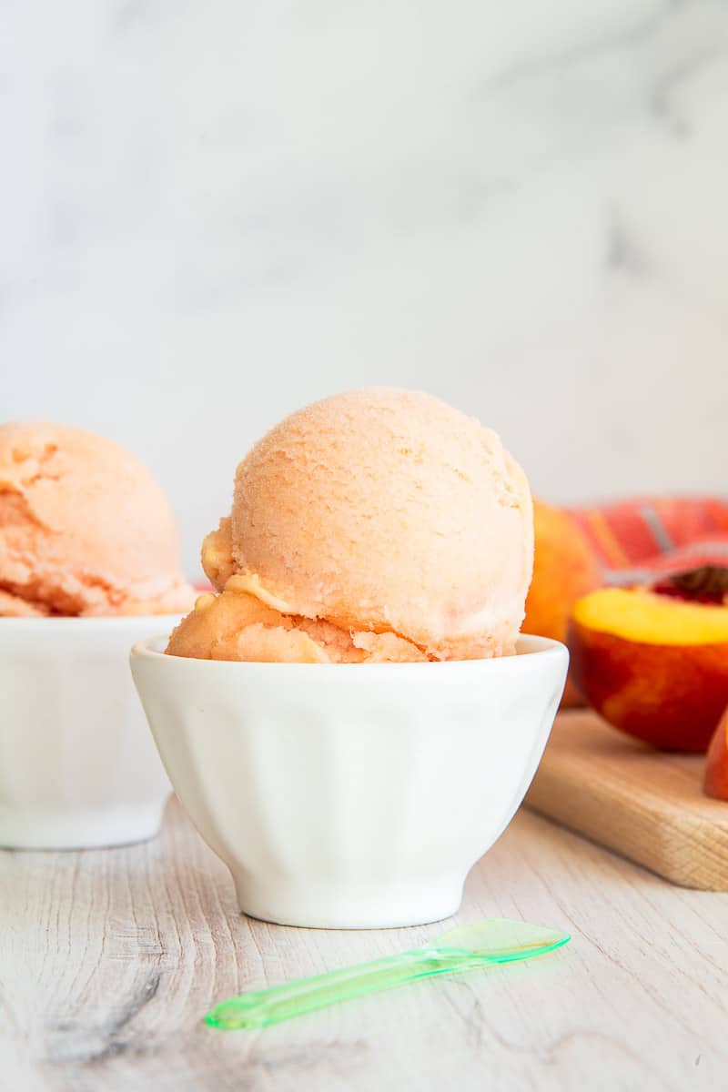 Peach Sorbet in white ceramic dessert bowls next to a wooden board with cut peaches on it.