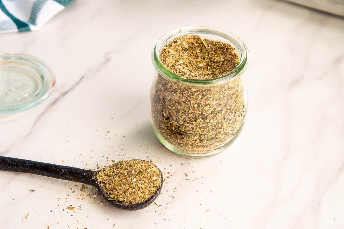 A jar of Greek Seasoning Blend next to a spoon filled with the seasoning blend.