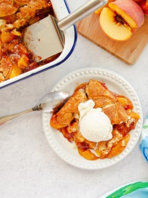 A serving of Homemade Peach Cobbler on a white plate topped with vanilla ice cream.