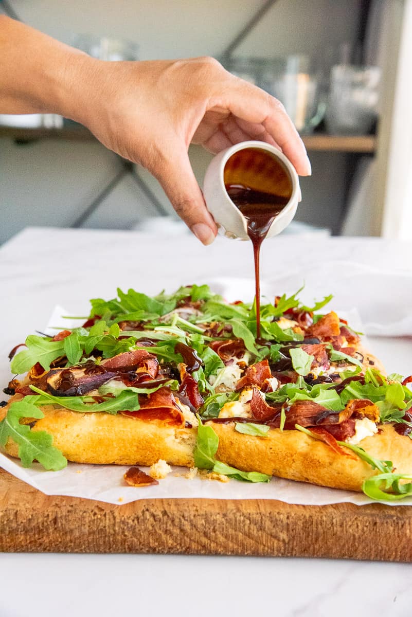 A hand pour a thickened balsamic vinegar from a small white pitcher over the flatbread.