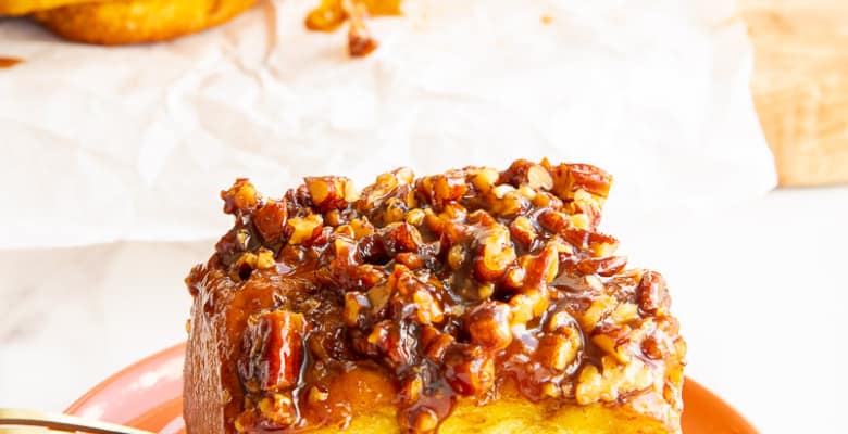 The Pumpkin Sticky Bun with its pecan caramel topping on an orange dessert plate in front of a board with the rest of the Pumpkin Sticky Buns with Pecans on it.