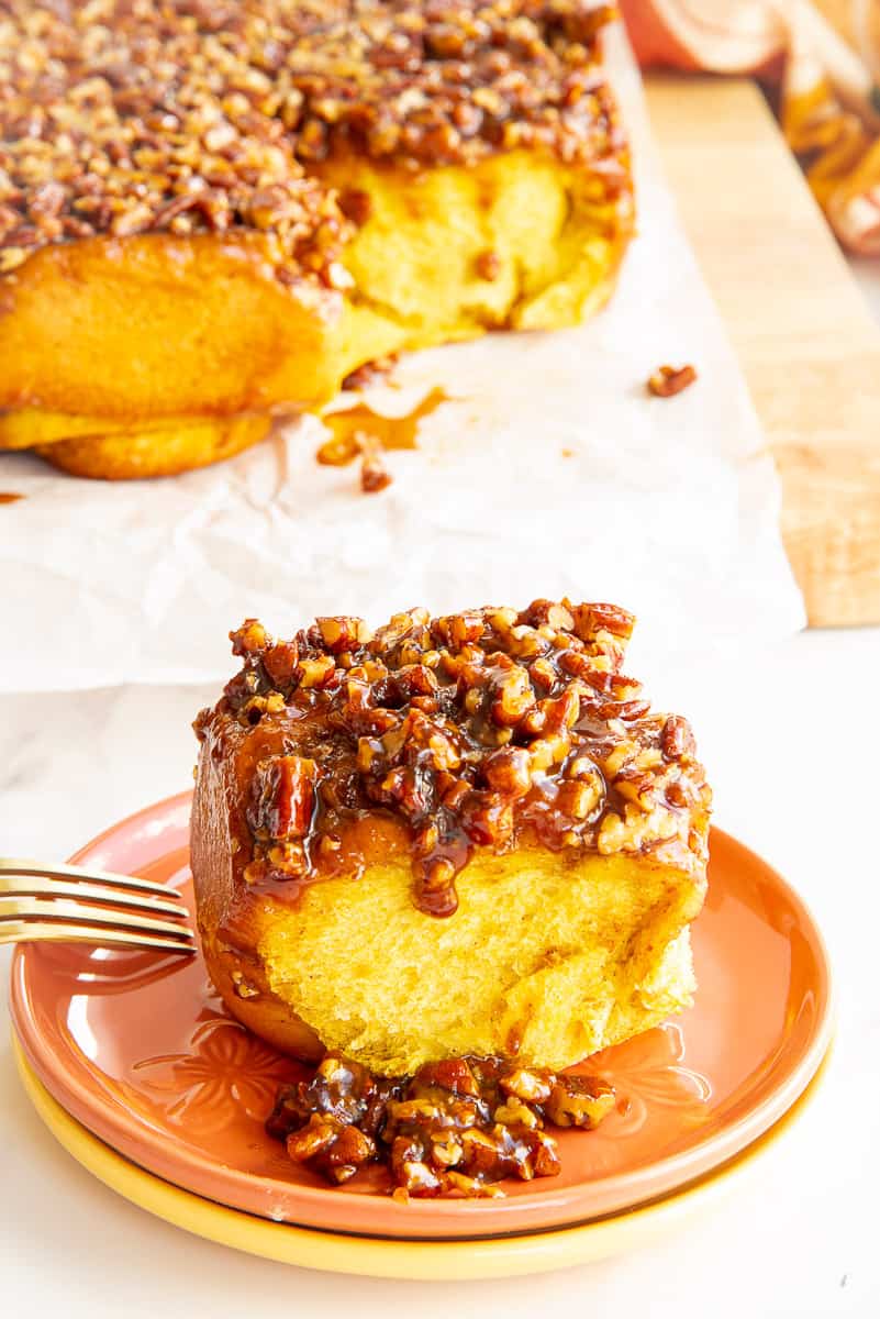 The Pumpkin Sticky Bun with its pecan caramel topping on an orange dessert plate in front of a board with the rest of the Pumpkin Sticky Buns with Pecans on it.