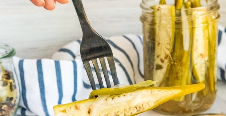 A hand uses a black fork to lift a Spicy Pickled Okra from a pile of okra on a small platter.