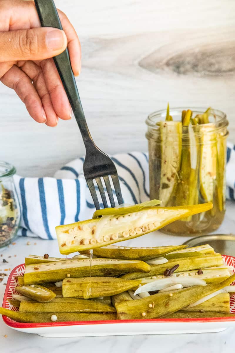 A hand lifts a Spicy Pickled Okra from piled of pickled okra on a red and white platter.
