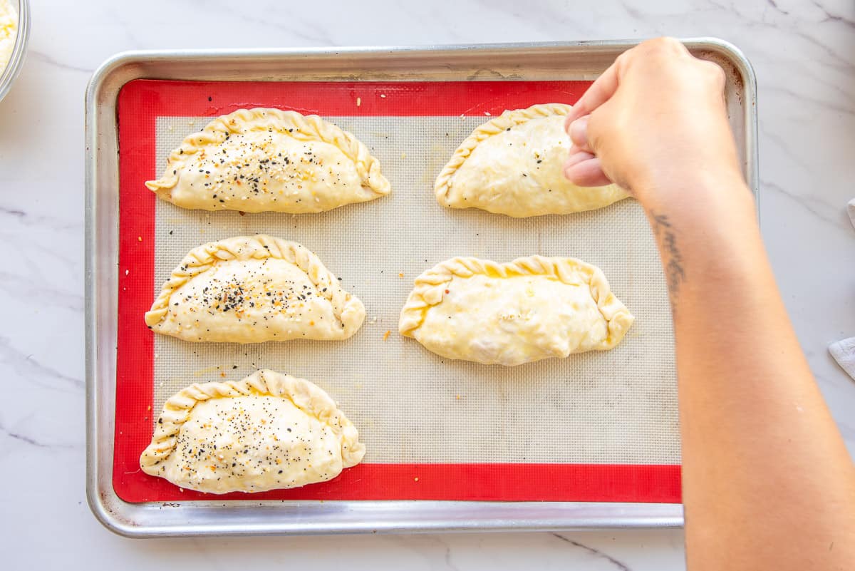 A hand sprinkles everything bagel seasoning over the surface of the unbaked pastries which are on a silicone mat-lined baking sheet.