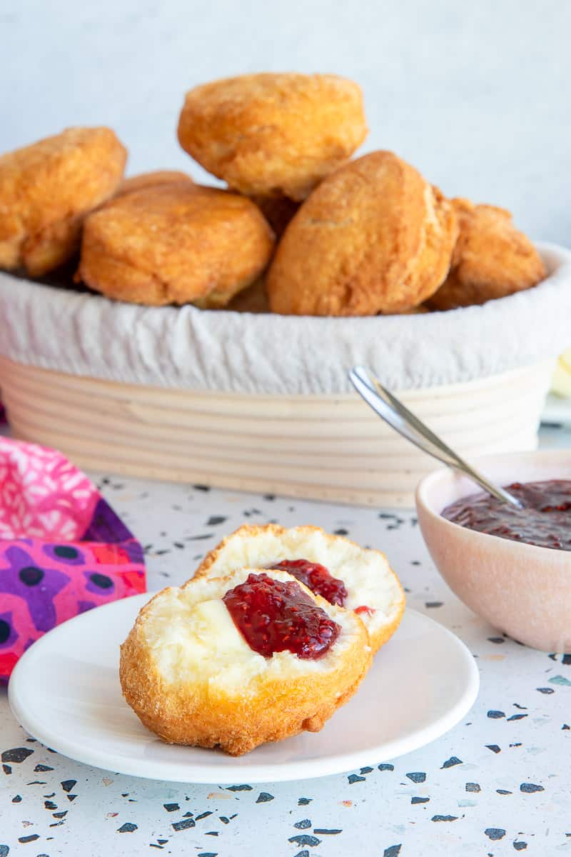 A Caribbean Johnny Cake opened on a white plate and spread with jam and butter. A basket of Johnny Cakes in the background.
