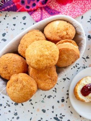 Caribbean Johnny Cakes in a bread basket next to a bowl of jam and a plate with a Johnny cake split in half, topped with butter and jam.