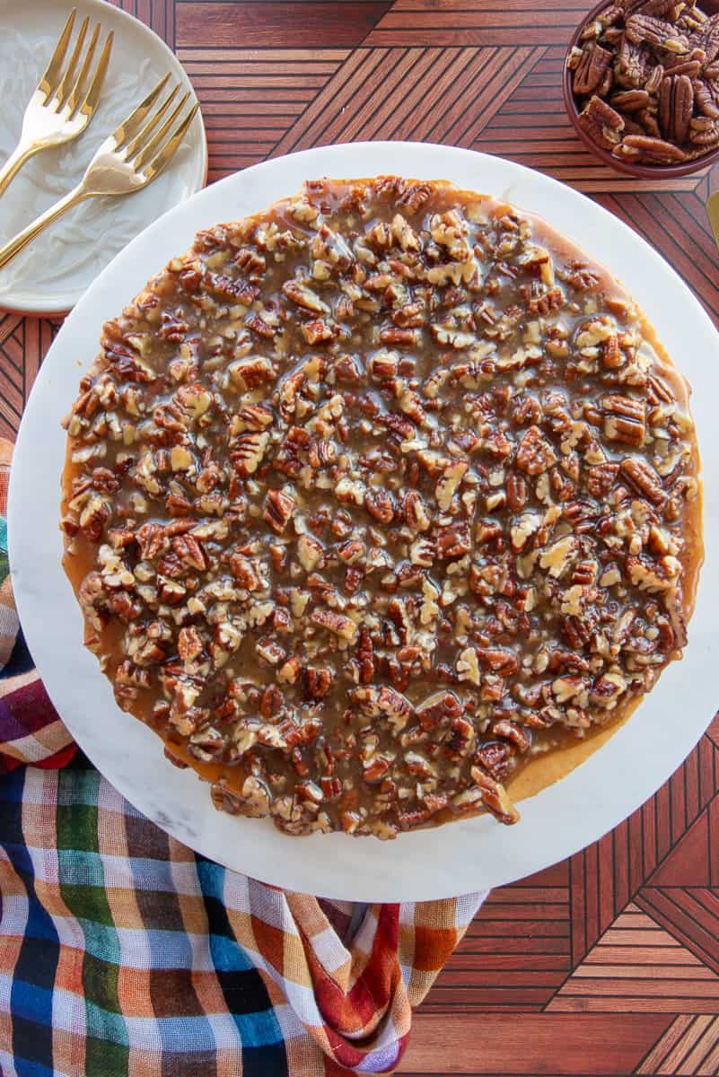 The cheesecake with the pecan praline topping on it next to a colorful kitchen towel and a bowlful of pecan halves.