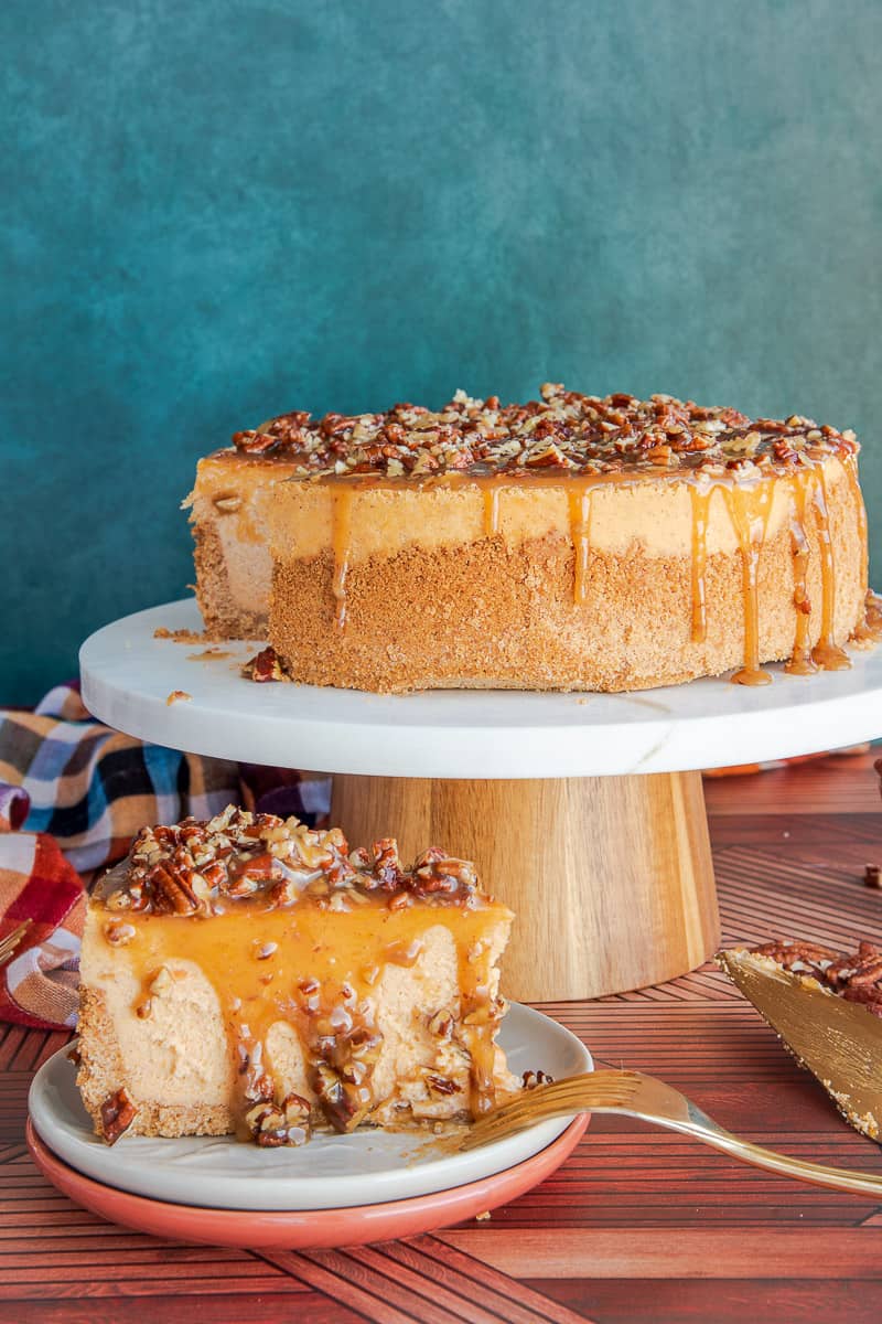 A slice of Sweet Potato Cheesecake with Pecan Praline Topping drizzling down the sides in front of a cake stand with the rest of the cheesecake on it.