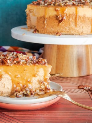 A single slice of Sweet Potato Cheesecake with Pecan Praline Topping on a stack of fall-colored dessert plates in front of a cake stand with the rest of the cheesecake on it.