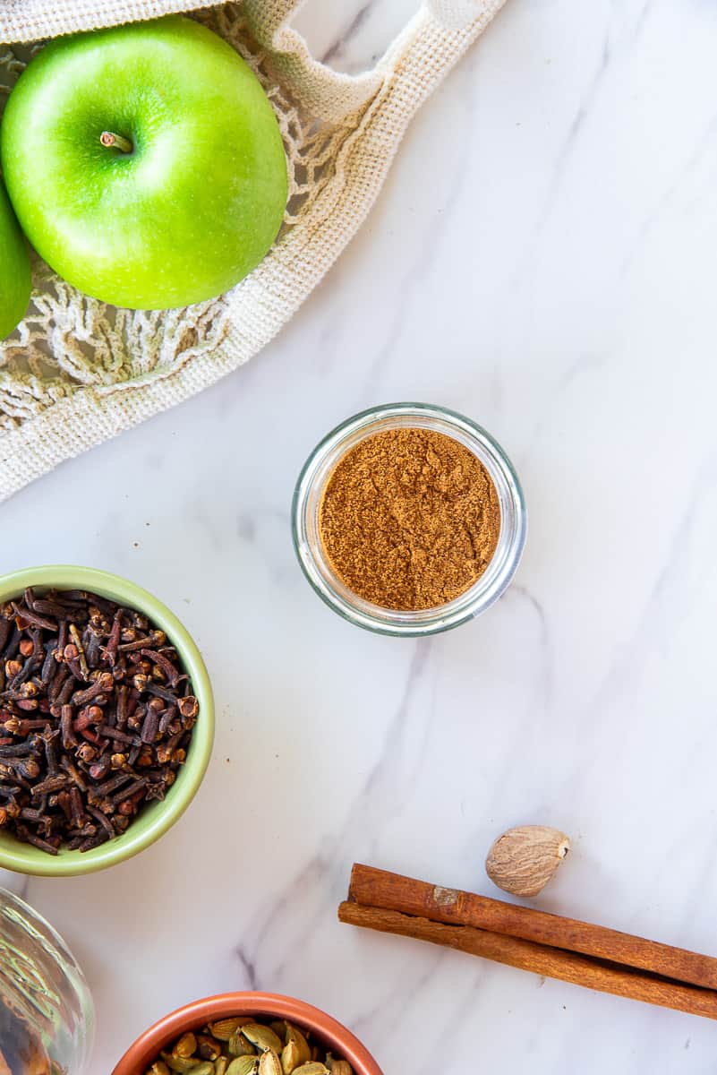 A jar filled with Apple Pie Spice Blend surrounded by green apples, a bowl of whole cloves, a cinnamon stick, nutmeg seed, and a bowl of green cardamom.