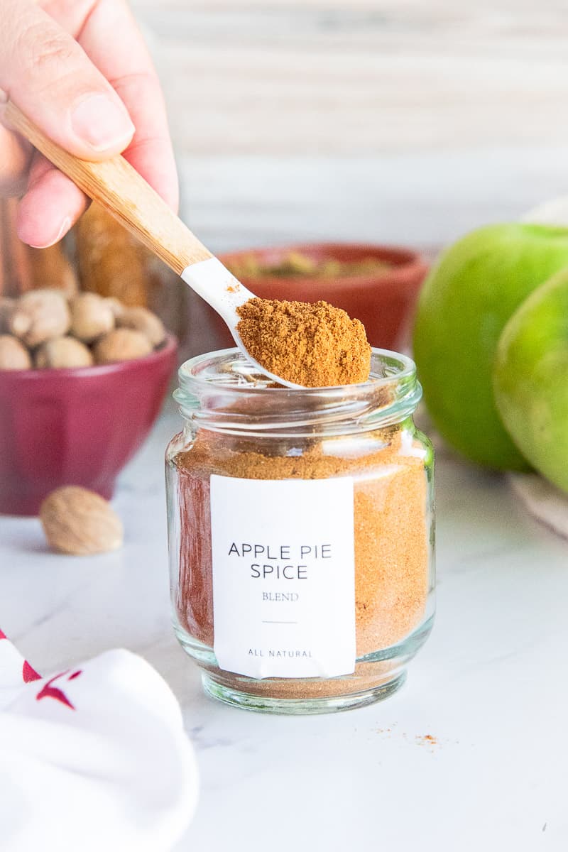 A hand lifts a spoonful of Apple Pie Spice Blend from its glass jar.