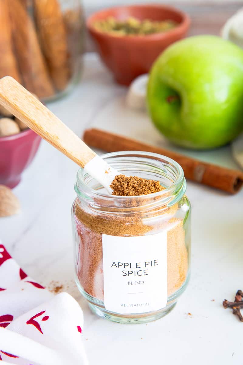 A spoon is inside a glass jar of the Apple Pie Spice Blend which is surrounded by the spices used to make it.