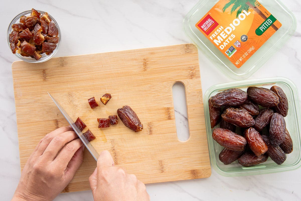 Hands hold a date and a chef's knife to cut the dates into small pieces on a bamboo cutting board.