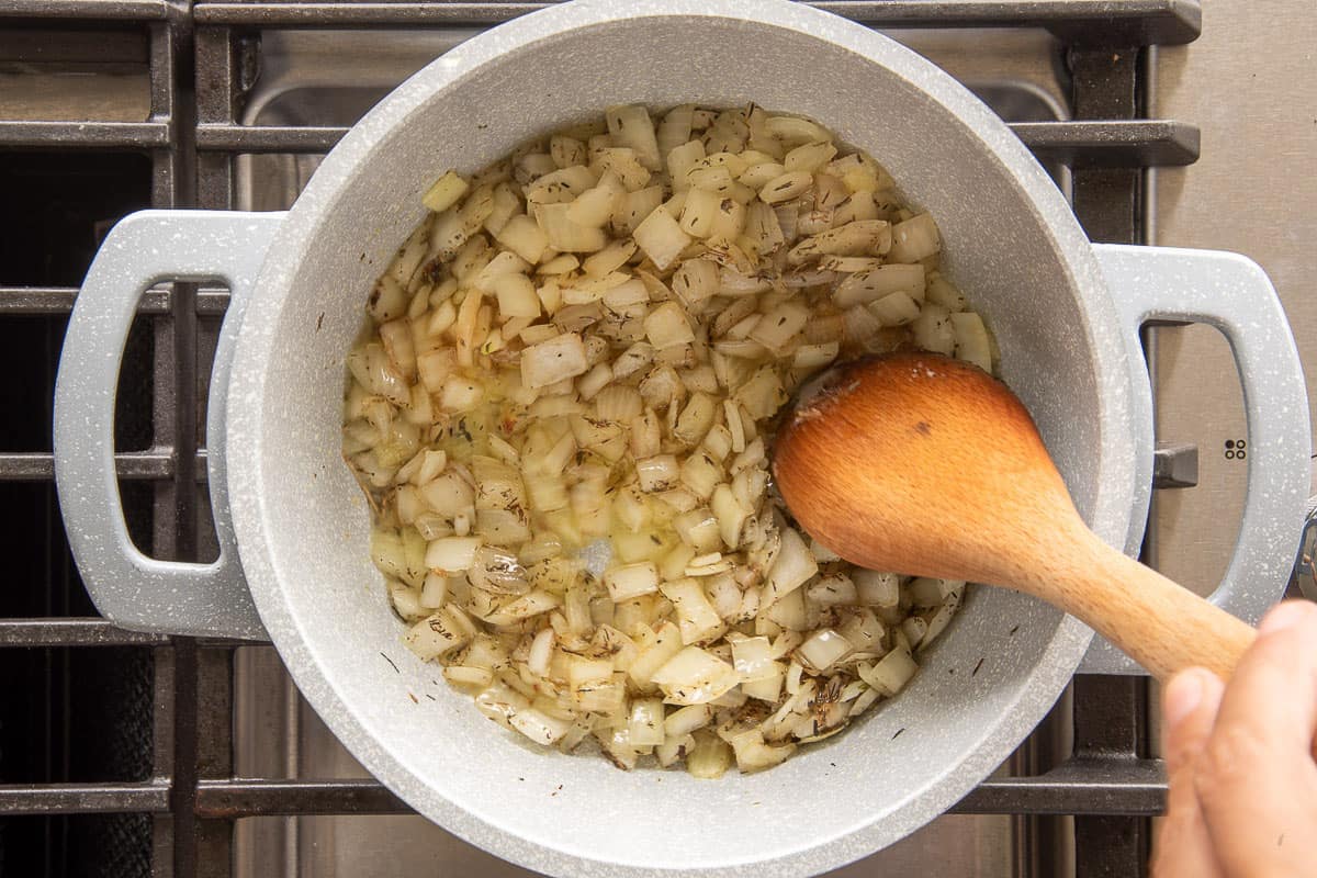 A wooden spoon stirs the brown sugar, spices, and thyme leaves into the sautéed onions in a pot.