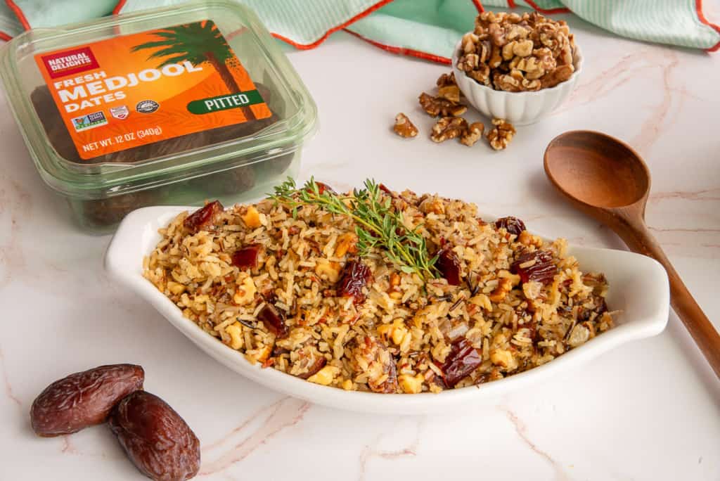 A white dish filled with rice pilaf mixed with chopped dates and nuts, garnished with herbs. Nearby are whole dates, a small bowl of chopped walnuts, a wooden spoon, and a container labeled "Fresh Medjool Dates." The setting is a white marble surface.