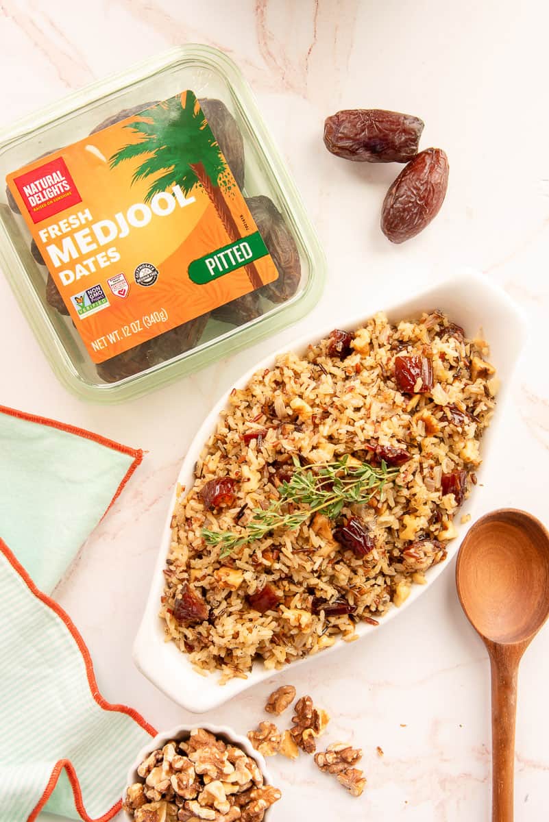A package of Natural Delights Medjool Dates next to a white serving bowl filled with Wild Grain Casserole with Caramelized Onion, Dates, and Walnuts and garnished with fresh thyme.