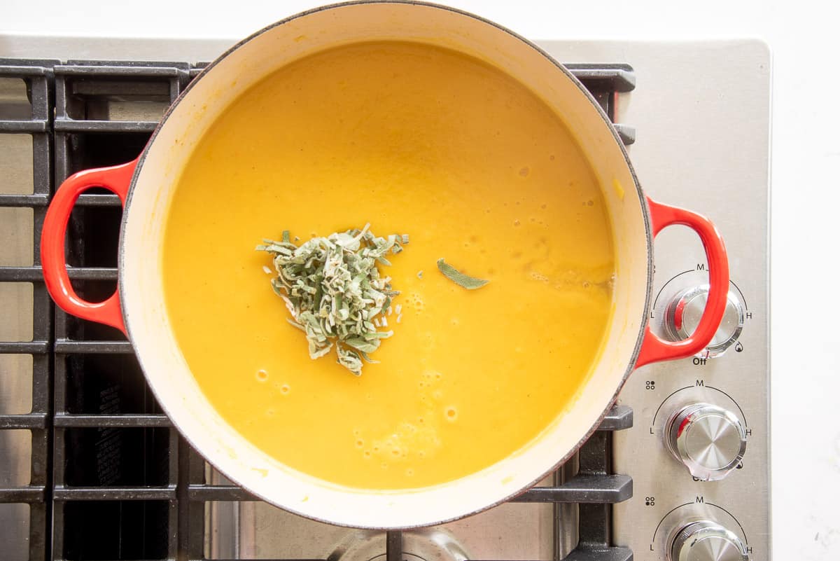 Fresh, chiffonade sage leaves are added to the Butternut Squash Soup in a red pot on the stove.