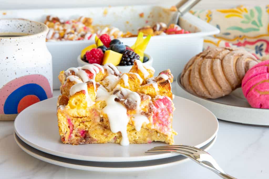 A serving of Concha Breakfast Bake on a white plate in front of a mug of coffee, a bowl of fresh fruit, and conchas.