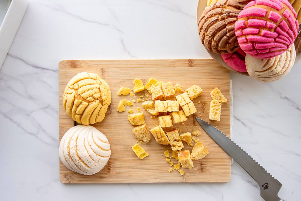 Multi-colored conchas are cut into cubes on a wooden cutting board with a serrated bread knife.