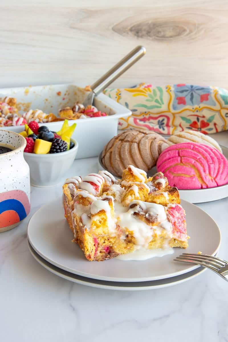A serving of Concha Breakfast Bake on a stack of two white plates in front of a plate with multi-colored mini conchas.