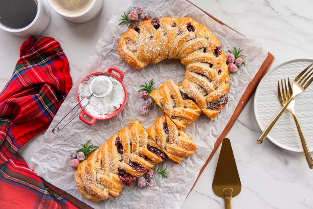 A beautifully braided pastry filled with berries and dusted with powdered sugar sits on parchment paper. It is accompanied by a small dish of powdered sugar, a red plaid napkin, gold cutlery, and plates on a marble surface.