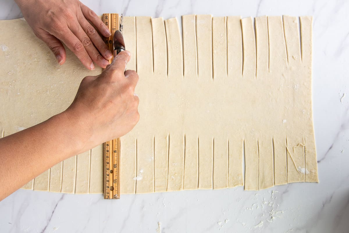 Hands use a ruler and pastry wheel to cut strips in the rectangle of dough.