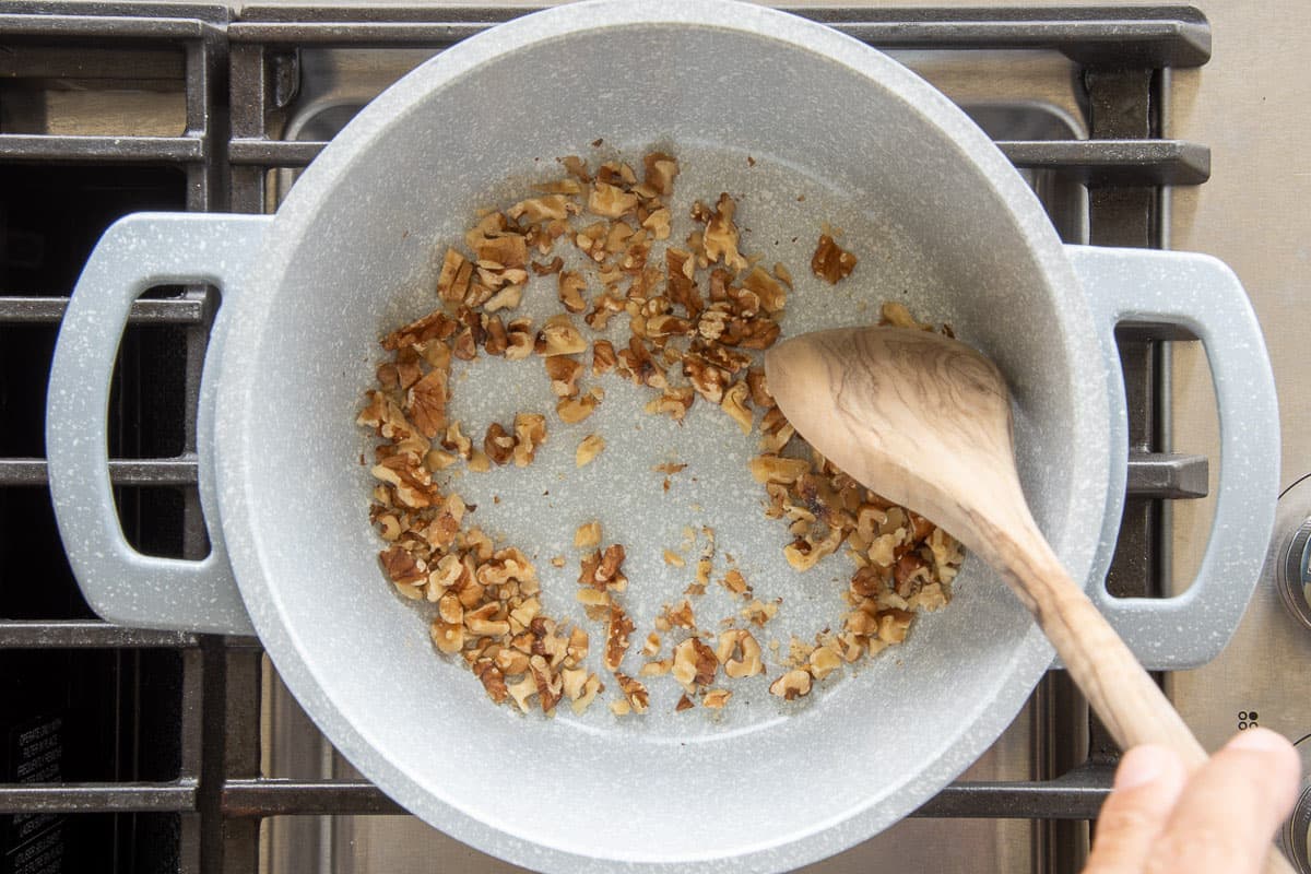 Chopped walnuts are stirred with a wooden spoon while being toasted in a pot on the stovetop.
