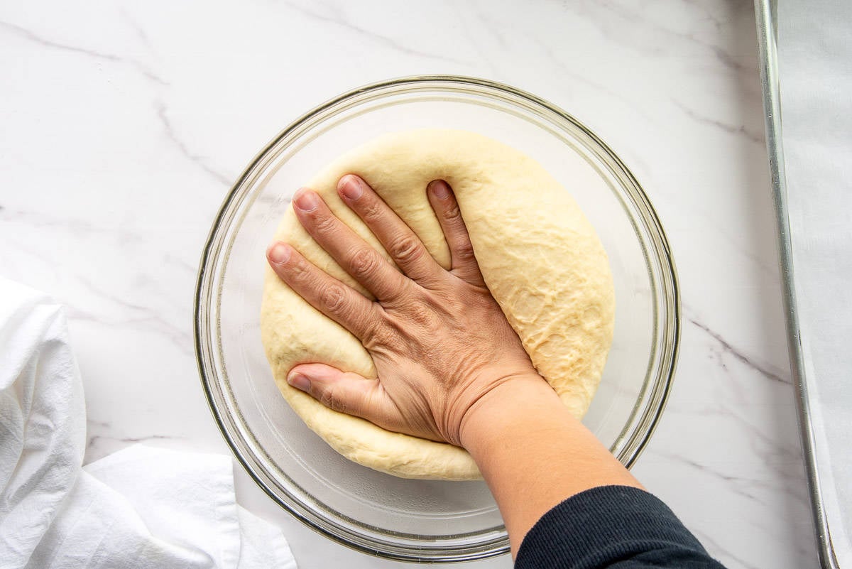 A hand pushes down the risen dough in a clear glass bowl to dispel the gases that have built up.