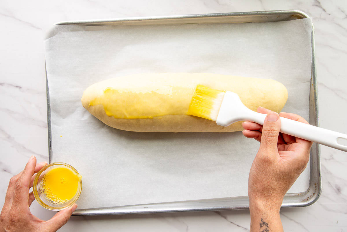 Hands hold a bowl of egg wash and brush the unbaked loaf with it using a white pastry brush.