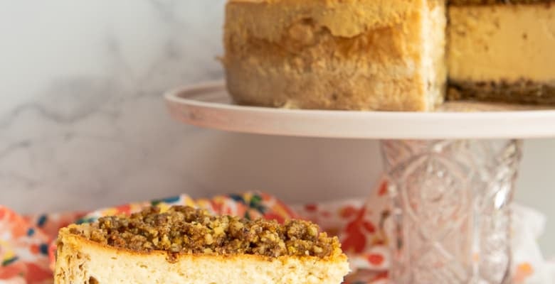 A full slice of Honey Baklava Cheesecake on a white plate in front of a cake stand with the rest of the cheesecake on it.