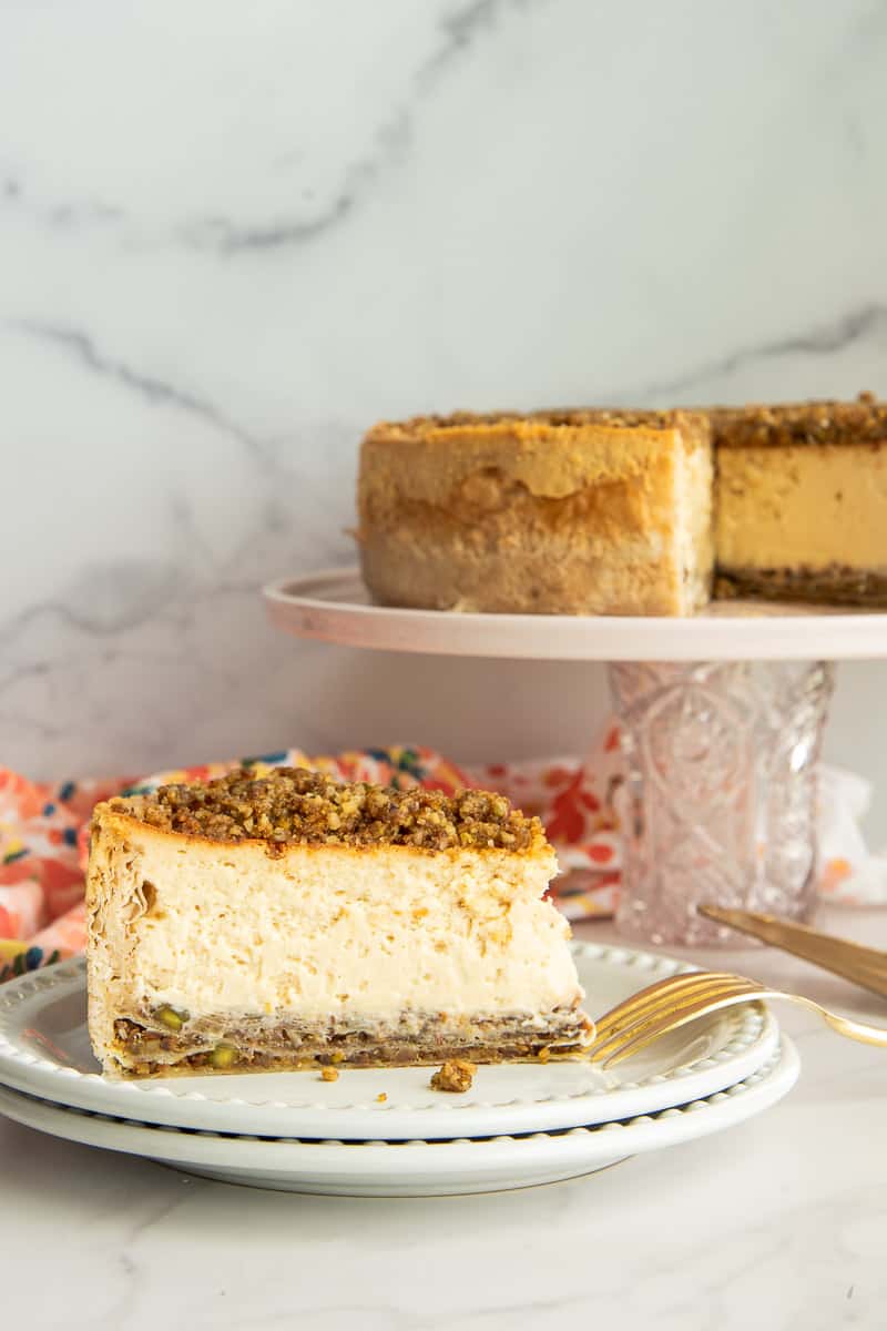 A full slice of Honey Baklava Cheesecake on a white plate in front of a cake stand with the rest of the cheesecake on it.