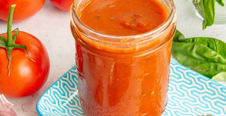 The Garlic Herb Marinara Sauce is in a glass jar that's sitting on a blue and white plate that has a spoon propped on it and is surrounded by the sauce's ingredients.