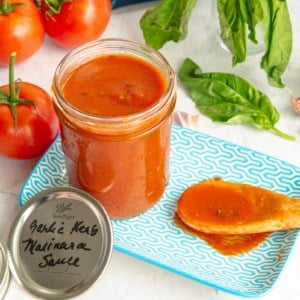A glass mason jar filled with Garlic Herb Marinara Sauce sits on a blue and white plate next to a wooden spoon with sauce on it and surrounded by the recipe's ingredients.