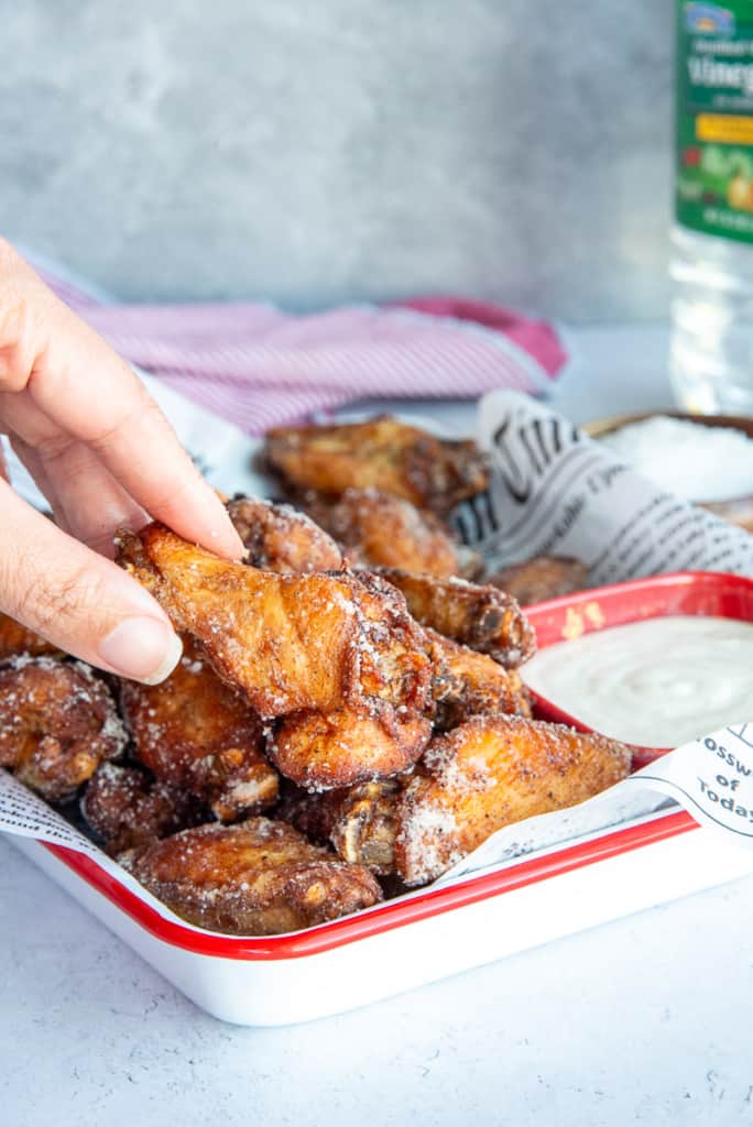 A hand picks up a drumette from a pile of Salt and Vinegar Chicken Wings on a white tray.