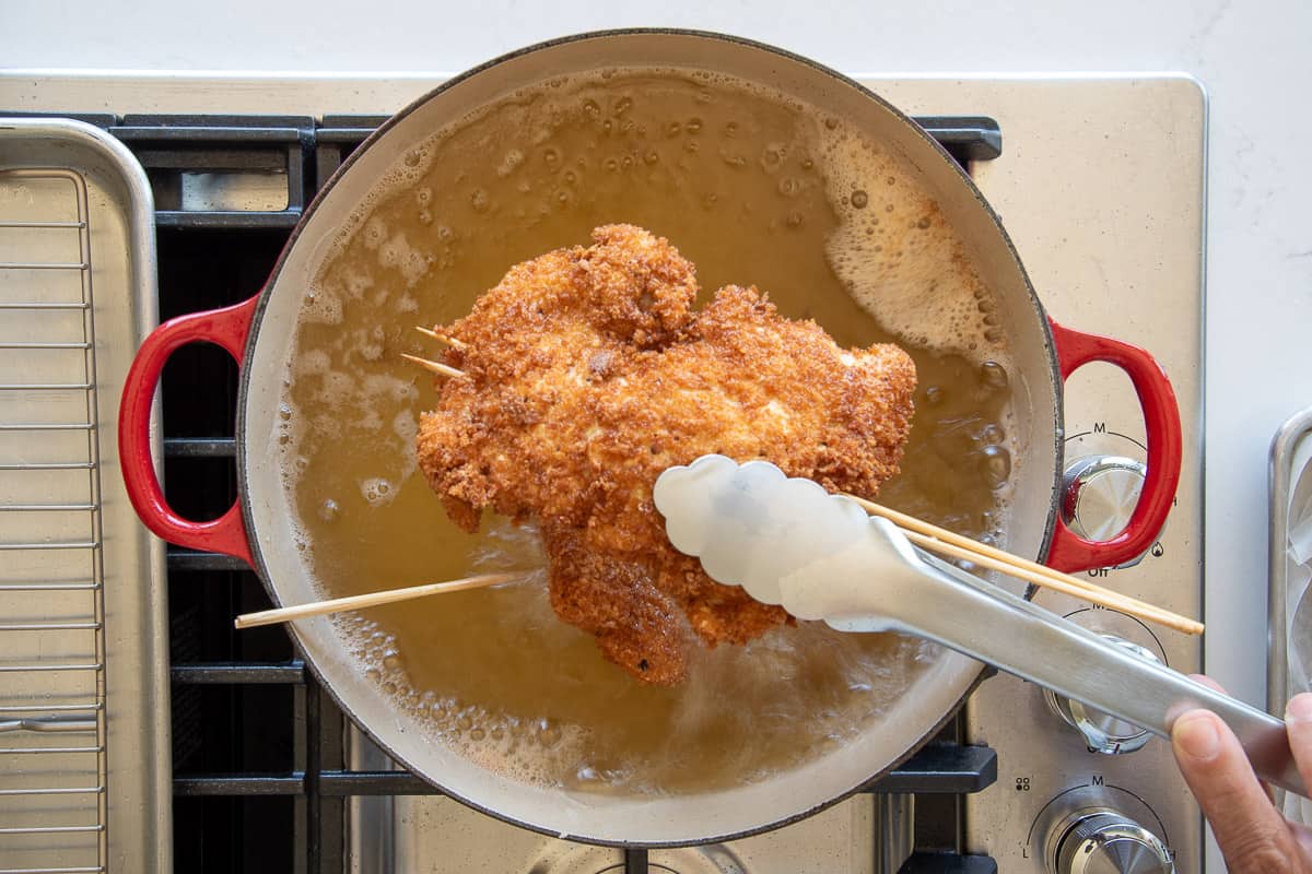 A fried thigh is lifted from the cooking oil with a pair of tongs.