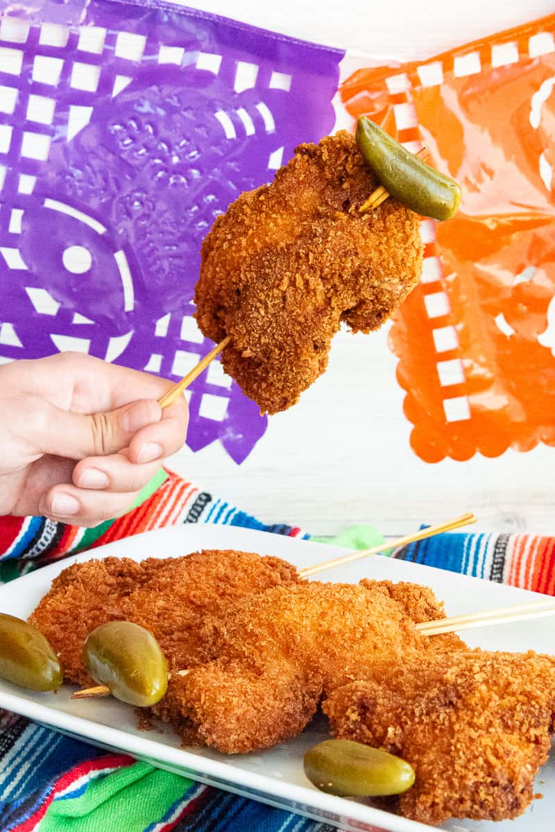 A hand holds a Chicken On A Stick topped with a pickled jalapeño pepper above a platter of Chicken On A Stick.